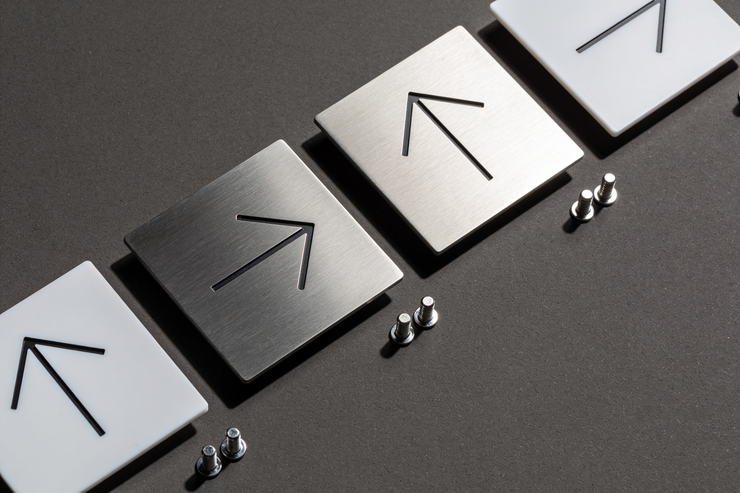 A row of brushed aluminium and acrylic wayfinding plates with directional arrows, laid out beside their standoff fixings.